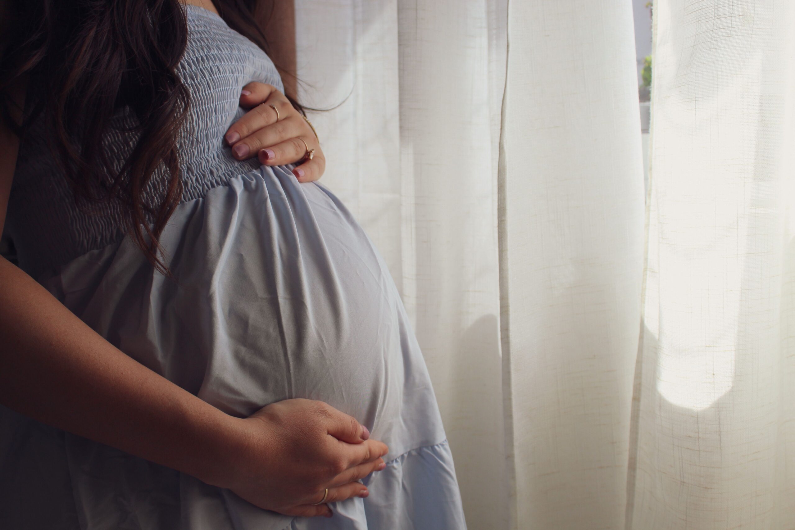 A peaceful moment of a pregnant woman cradling her belly by a sunlit window indoors.