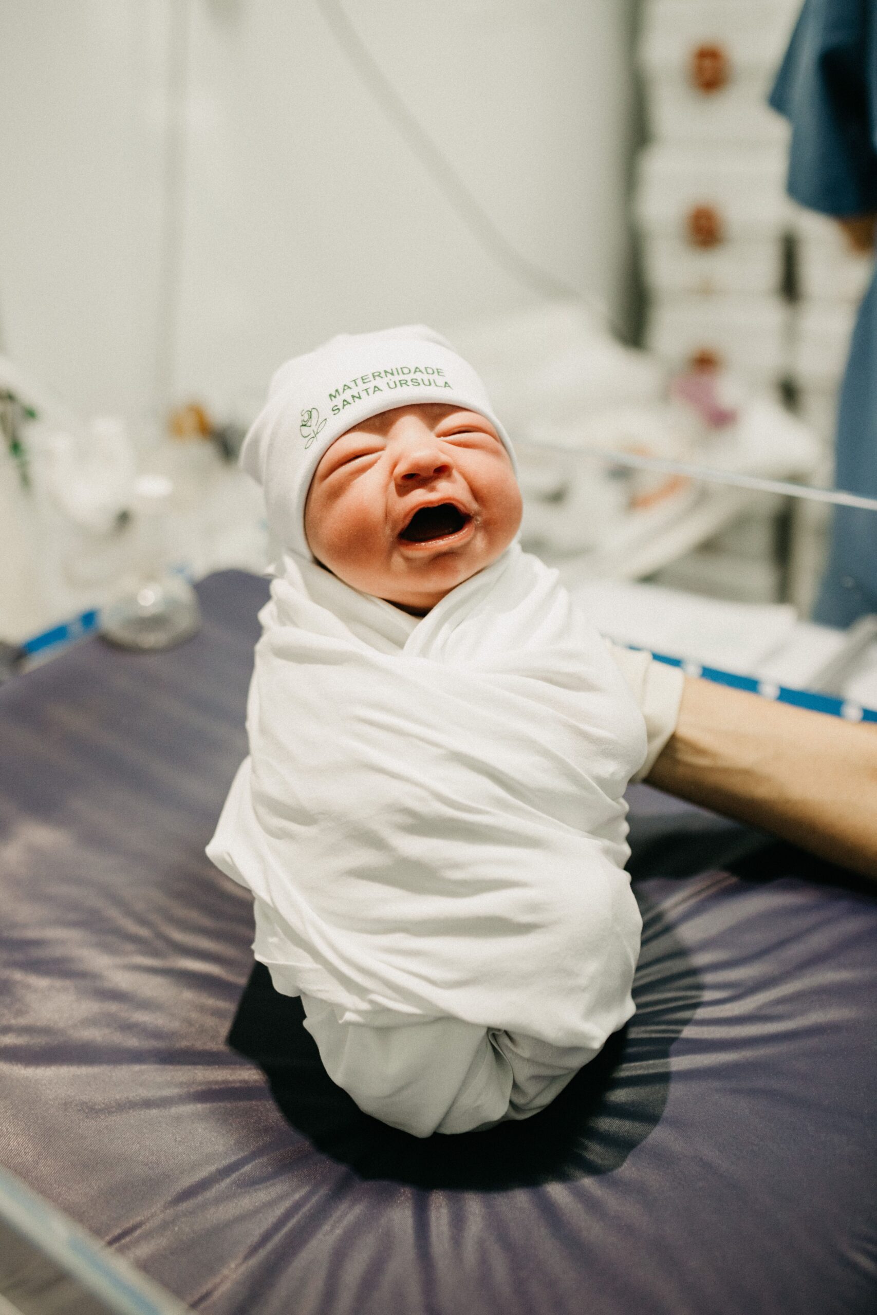 A cute newborn baby wrapped in a white blanket, crying in a hospital setting.