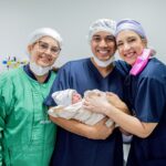 Smiling healthcare professionals holding a newborn baby in a hospital room.