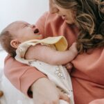 Side view of crop anonymous mother consoling crying infant baby in arms while sitting in light room near wall at home