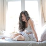 Woman sitting on bed holding her stomach, showing signs of discomfort or pain indoors.