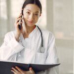 Crop ethnic female doctor in medical uniform with stethoscope and notebook talking on cellphone while standing in clinic corridor