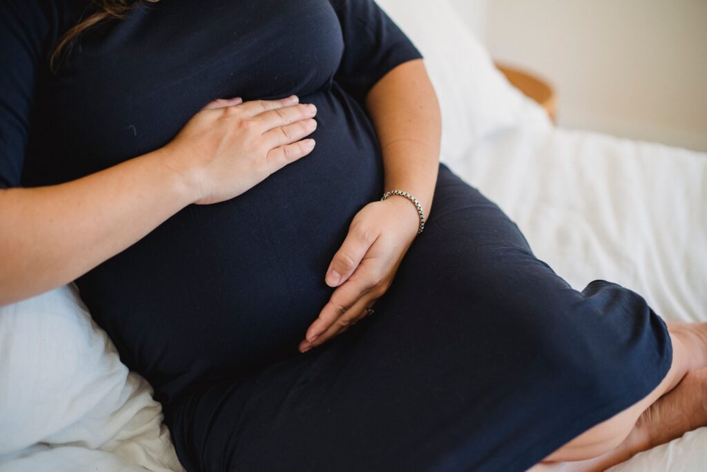 High angle of crop anonymous expectant female in dress stroking tummy while resting on soft bed in house