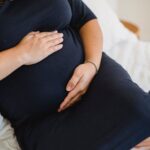 High angle of crop anonymous expectant female in dress stroking tummy while resting on soft bed in house