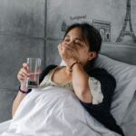 Elderly sick Asian female lying in comfortable bed and drinking pill with glass of water