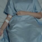 A patient lies in bed wearing a hospital gown with an IV drip attached, evoking a sense of illness.