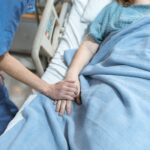 A nurse in scrubs offers comfort by holding a patient's hand in a hospital bed.