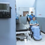 A nurse helps a patient in a wheelchair down a hospital corridor, reflecting care and medical professionalism.