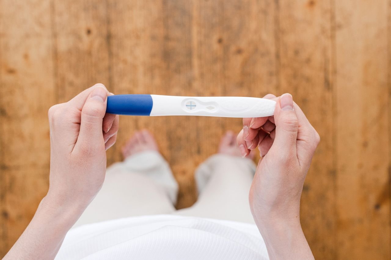 Top view of anonymous barefoot female with pregnancy test in hands standing on wooden floor in light room at home