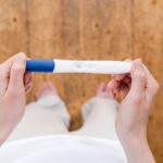 Top view of anonymous barefoot female with pregnancy test in hands standing on wooden floor in light room at home